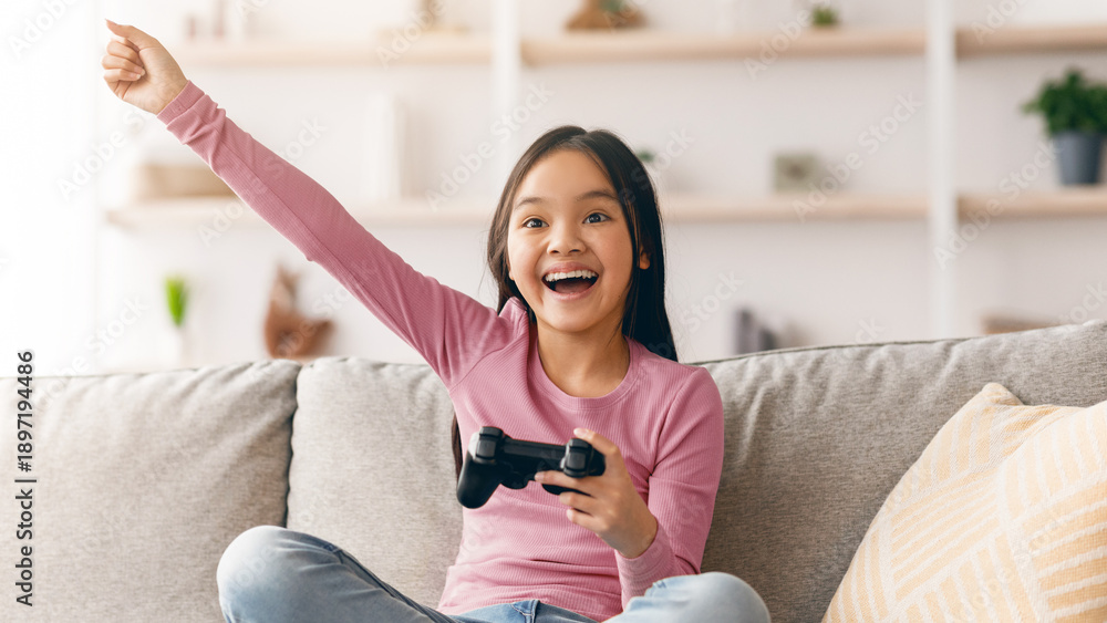 Fototapeta premium A young girl sits on a couch in a living room, holding a game controller and smiling broadly. She raises one arm in celebration, showing happiness after a gaming achievement.