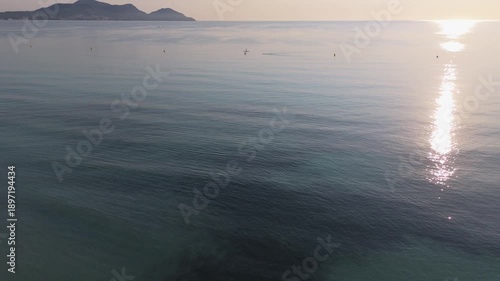Calm waters of Playa de Muro beach in Mallorca, Spain, with paddleboarder gliding across the surface under the warm sunlight and distant mountains