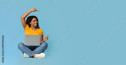 Happy young black woman sitting on floor with brand new laptop, pointing and looking at copy space over blue studio background, pretty african american lady using notebook, panorama