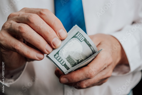 man is counting multiple hundred dollar bills in their hands. individual is in business environment, wearing white shirt and blue tie. focus is on cash they hold. close up.