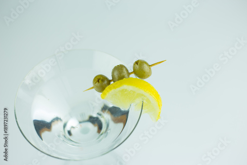 A close up, overhead view of a classic martini with a slice of lemon and three green olives against a white background.