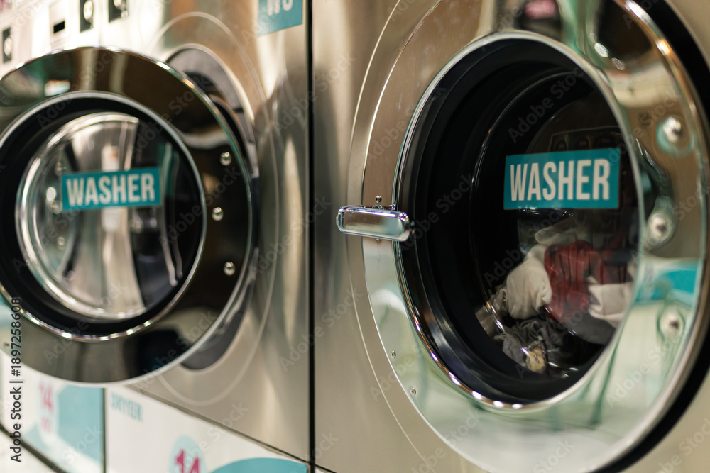 Obraz premium In a public laundry, several washing machines are working. Each machine has clothes inside them. People are nearby, waiting for their laundry to finish. It is daytime