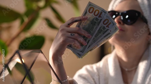 Confident young woman in a bathrobe and sunglasses proudly fanning herself with a wad of hundred-dollar bills. Concept of wealth and luxury