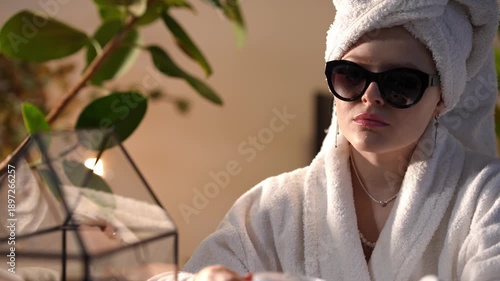 Young woman in a white bathrobe and towel on her head enjoying the morning sun. She is elegantly drinking coffee and looking at the camera