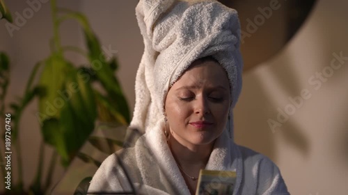 Young woman in a bathrobe with a towel on her head kissing a large wad of cash, expressing joy and greed in a bright room