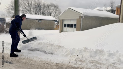 Adult man shoveling heavy snow outside a garage in a residential alley after a winter storm, clearing a driveway and snowbank in cold conditions. Pittsburgh suburbs. 15034