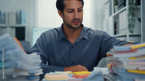 Overworked young Latin businessman reading document in large stack of paperwork files at office desk. Tax audit deadline, accounting stress, corporate burnout, bookkeeping, bureaucracy concept.