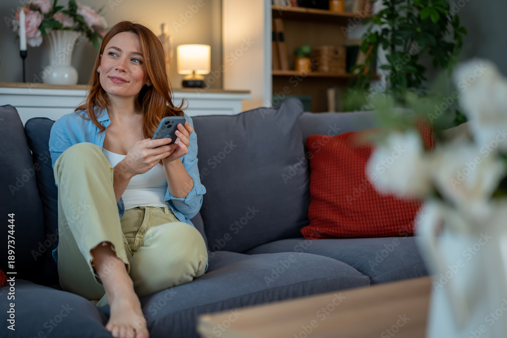 Obraz premium Redhead woman relaxing on sofa holding smartphone looking away