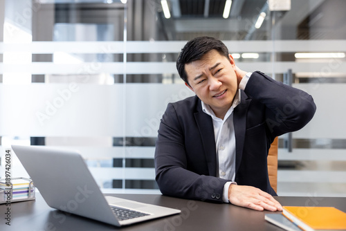 Asian businessman suffering from acute neck pain and discomfort, touching his sore neck while working on a laptop at his desk in a contemporary office setting, experiencing stress and health issues