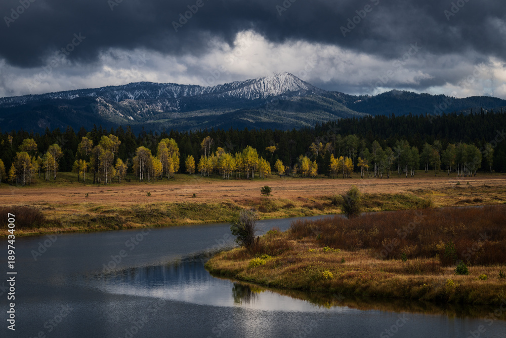 Fototapeta premium Breathtaking landscape featuring a winding river, golden fall foliage, and a snow-capped mountain under dramatic storm clouds. Perfect for travel and environmental themes.