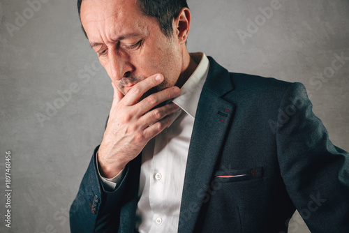 Middle-aged businessman in dark suit jacket, white shirt and tie, hand thoughtfully on chin while gazing downward with visible worry and tension.