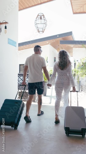 Rear view of a man and woman holding hands while walking with their luggage. The couple is arriving at a sunny tropical hotel for vacation. Vertical