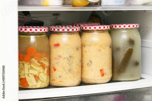 Homemade preserved soups in glass jars. Chicken broth and Thai soup stored on fridge shelf. Traditional pickled cucumbers in brine. Home food preservation. Ready meals prepared in advance.