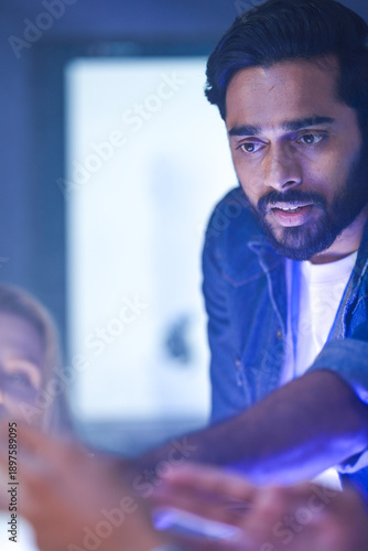 An Indian man holds a glowing robotic arm prototype in a dark tech lab. He is presenting the futuristic hand to a colleague during a late night innovation meeting.