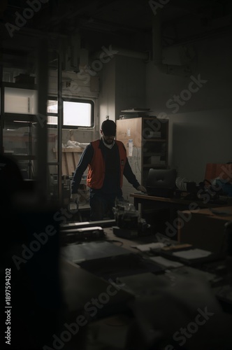 Worker in Safety Vest Moving Through a Dimly Lit Workplace With Tools and Desks