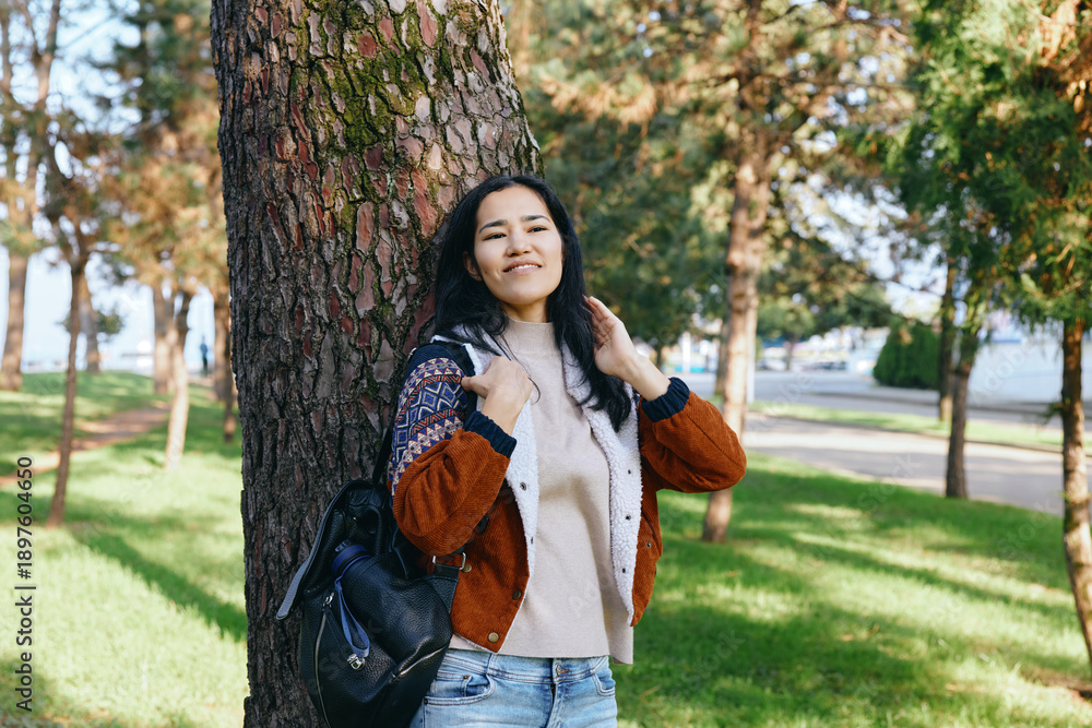 Fototapeta premium Woman leaning against a tree in a park, autumn sunlight and nature setting, casual sweater and backpack for outdoor lifestyle and travel imagery