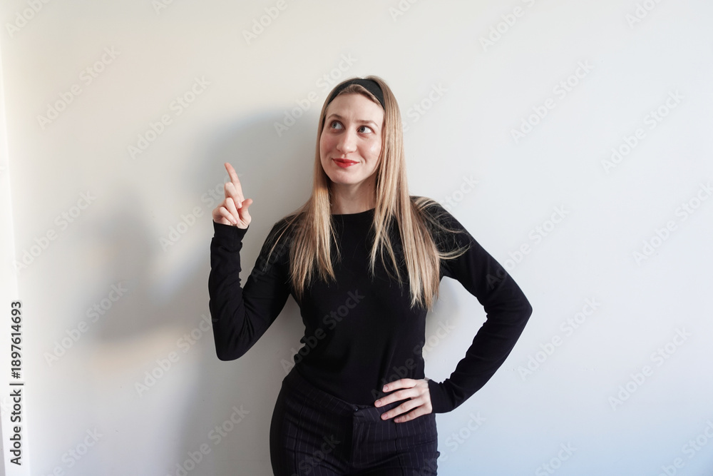 Fototapeta premium Młoda kobieta w czarnym stroju wskazuje palcem w górę i patrzy w bok na jasnym tle. Koncepcja pomysłu, inspiracji i komunikacji wizualnej.