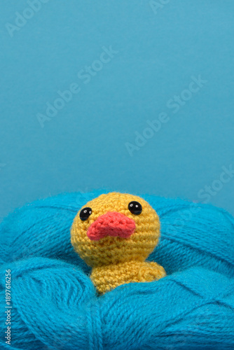 A small yellow crocheted duckling toy sits nestled in a large ball of blue wool yarn against a matching blue background with copy space above