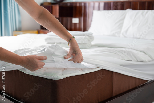 The hands of a person tucking a white sheet onto the edge of a mattress while making a bed. Hotel room cleaning