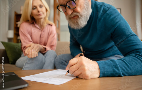 Senior Couple Filling In Form Signing Papers Sitting On Sofa Near Table Indoors. Paperwork Concept. Elderly Man Writing His Will In Notary Office. Cropped Shot, Selective Focus