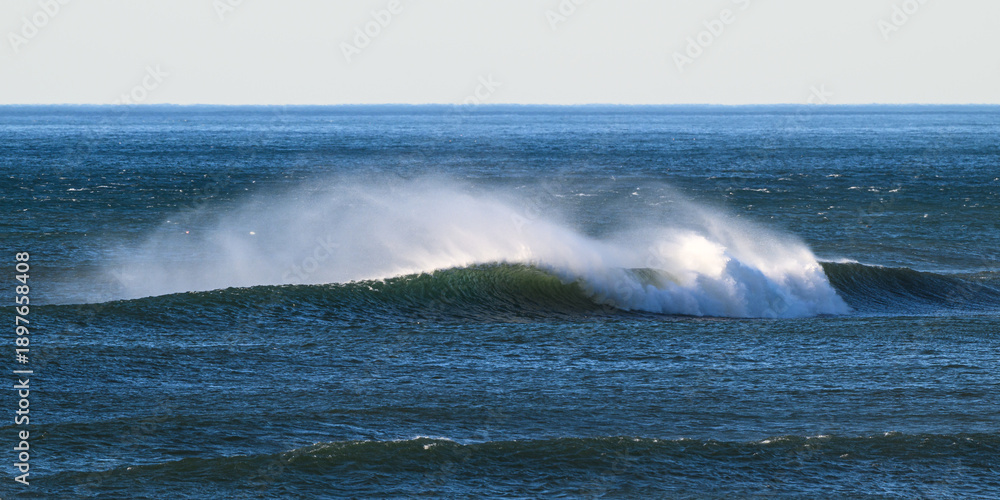 Naklejka premium Ocean wave blown by strong wind pulling spray away from the crest towards horizon