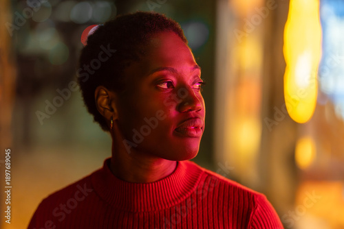 Young woman with short Afro hair at night, looking away with calm expression, warm neon light on face, relaxed posture against blurred background