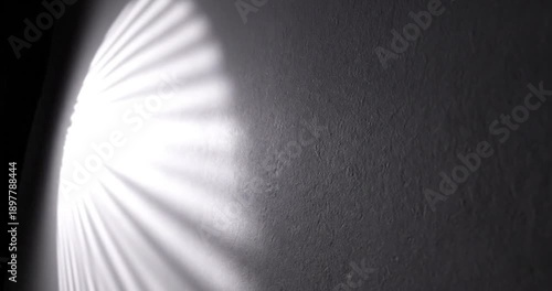An abstract and captivating play of light and shadow dynamically interacts with a textured wall surface. Brilliant rays of light, fanned out in a striking pattern, slowly sweep across the dark, rugged