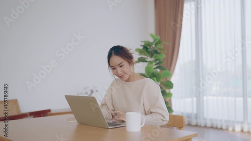 Young Asian woman using laptop computer for business studying, having video call with online virtual webinar training meeting, writing notes. Focused student female working in living room at home