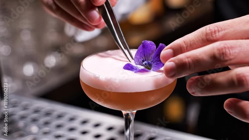 Bartender hands meticulously garnish a craft cocktail with a vibrant purple flower. The elegant drink features frothy pink foam and a rich amber liquid.