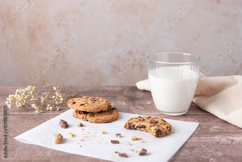 Cuadro en lienzo Chocolate chip cookies with a bite and crumbs on white paper next to a glass of milk, beige background and wooden table in horizontal composition with copy space