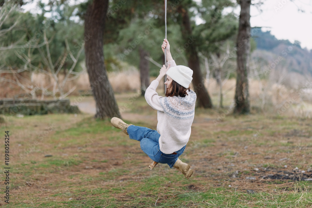 Fototapeta premium Girl swinging on a rope swing in a park, wearing a knit sweater, hat and boots, mid motion with trees and grass in the background, capturing carefree outdoor play in nature