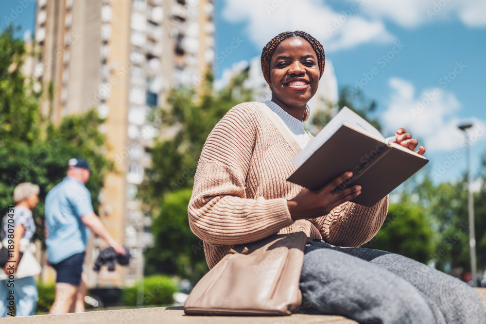 Fototapeta premium Cheerful African American woman relaxing in the city park, reading a book.