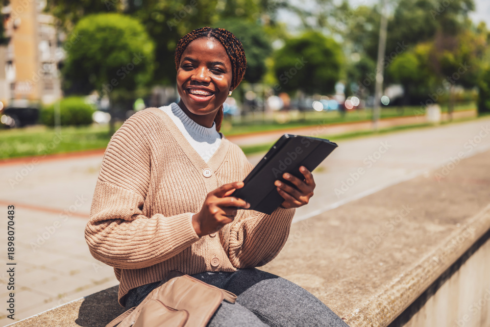 Obraz premium Portrait of a joyful Black female using a digital tablet for online communication. She relaxes outdoors after work.