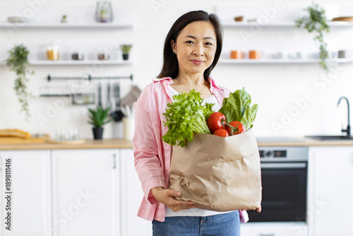 An Asian woman smiles while holding a bag of fresh produce in her bright, modern kitchen