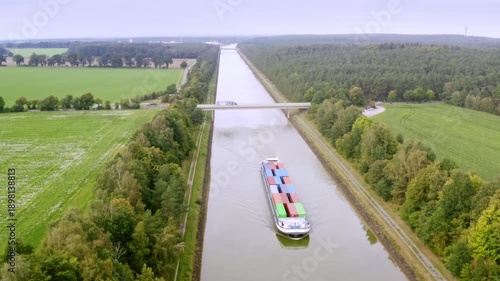 Aerial view of a long container ship navigating a narrow canal. It is passing under a bridge crossed by a truck, surrounded by a lush landscape of green forests and open fields.