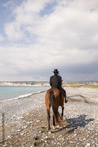 The power of women. Rear view of a woman riding a brown horse on a beach near the sea. Equestrian lifestyle. Girl in a black suit and a black hat with a veil.