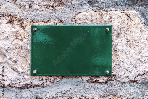 Blank green metal plate mounted on a textured stone wall with bolts. Empty signage mockup on old rock background for custom text or house numbers in Europe