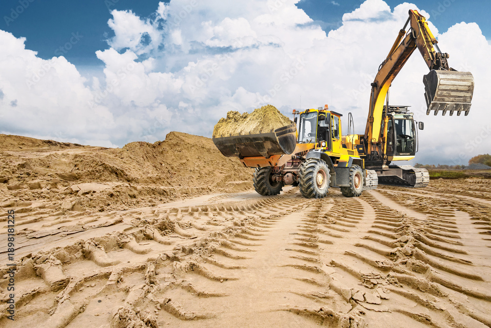 Obraz premium Heavy machinery moves through sandy ground making deep tracks while a loader and excavator work in the background on a cloudy day at a construction site