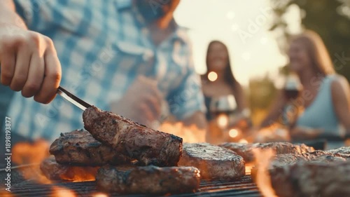 Close-up of grilling steaks, with a person handling tongs, and blurred figures of people in the background
