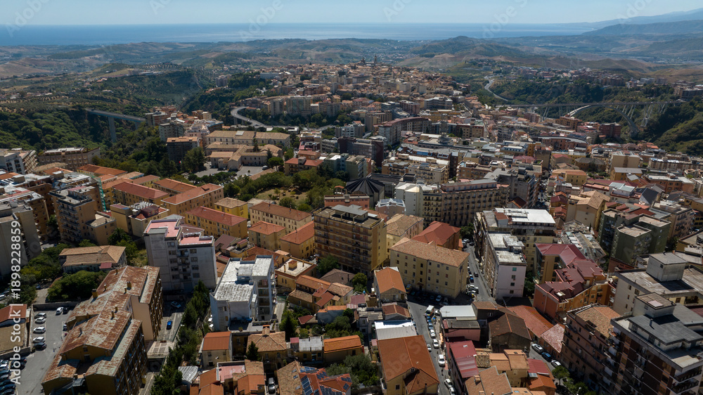 Fototapeta premium Panoramic aerial view of the city of Catanzaro, capital of the Calabria region in southern Italy. In the foreground are the town's houses, buildings, and rooftops. The Ionian Sea is on the horizon.