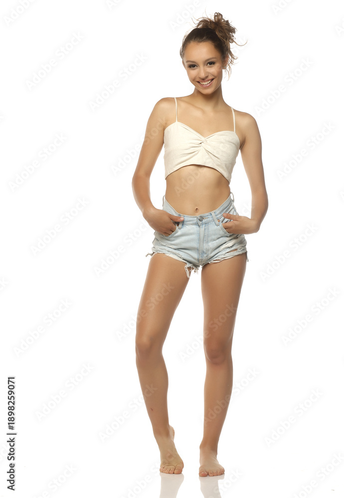 Fototapeta premium Studio shot of a smiling young woman with brown hair wearing a beige crop top and denim shorts standing full body on a white background.