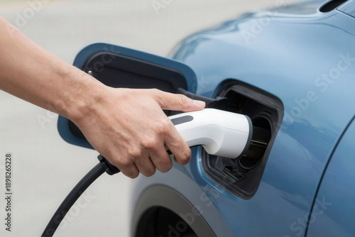 Charge. Hand holding electric vehicle charging connector while plugging it into the charging port of a blue electric car, showcasing the modern technology in an outdoor setting