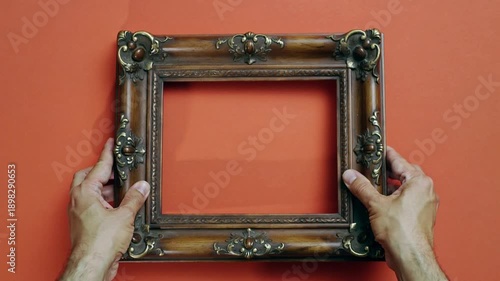 Ornate Wooden Frame with Bronze Accents Held by Hands on Orange Background