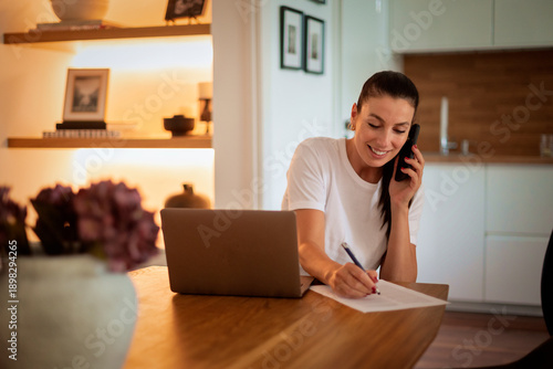 Confident woman sitting at table, using computer and holding a cell phone in his hand and talking on the phone