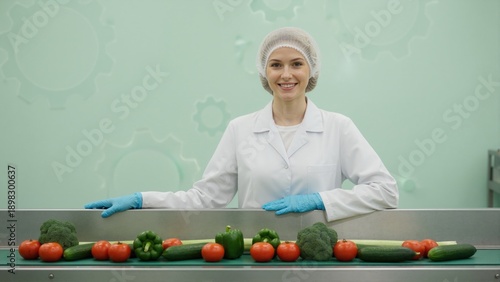 Smiling female worker at food processing facility. Professional woman in hairnet and lab coat with fresh vegetables on conveyor belt. Food safety and quality control concept