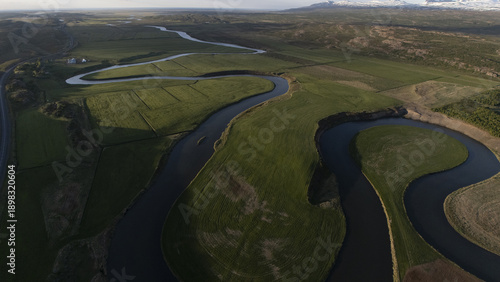 Aerial view of the serene, winding river cutting through lush green fields, casting shadows and reflecting the sky above, Selfoss, Iceland.