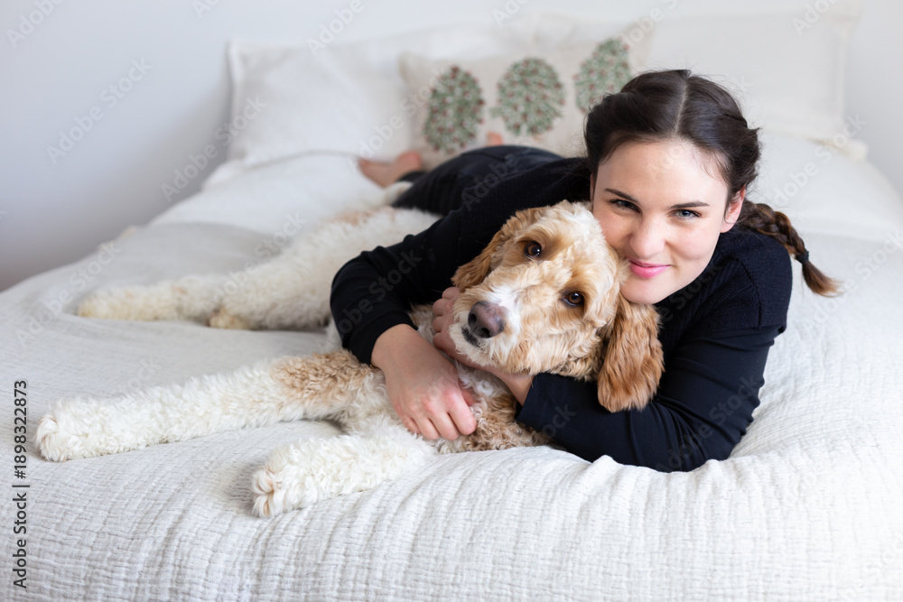 Obraz premium Cute brunette smiling young woman lying down on bed hugging adorable friendly Parti Goldendoodle with white and ginger coat