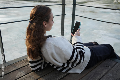 Teenage girl sitting in city park and smiling while holding smartphone. Modern youth lifestyle, quiet moment of balance and pause in everyday life, third place concept, mindful screen time