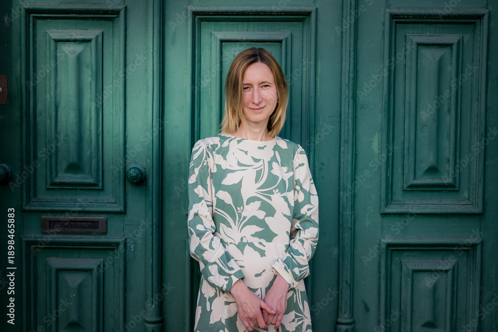 Fototapeta premium A woman stands in front of a large green wooden door, wearing a light patterned dress. She looks calmly at the camera in soft daylight, with a simple urban background.