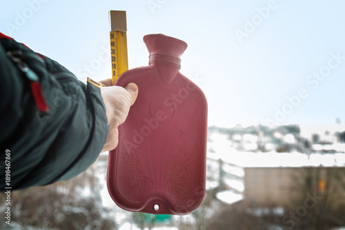 Obraz na płótnie Hand in a warm jacket holding an outdoor thermometer and a rubber hot water bottle against a winter landscape background, close-up